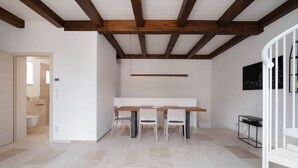 Een modern interieur met een houten eettafel en stoelen, een wit plafond met zichtbare balken, en een betegelde vloer.