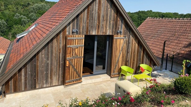 Een houten schuur in de stijl van een boerderij, met een schuin dak, een grote houten deur en een terras met stoelen.