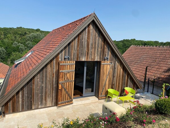 A wooden barn-style building with a sloped roof, a large wooden door, and a patio area with chairs.