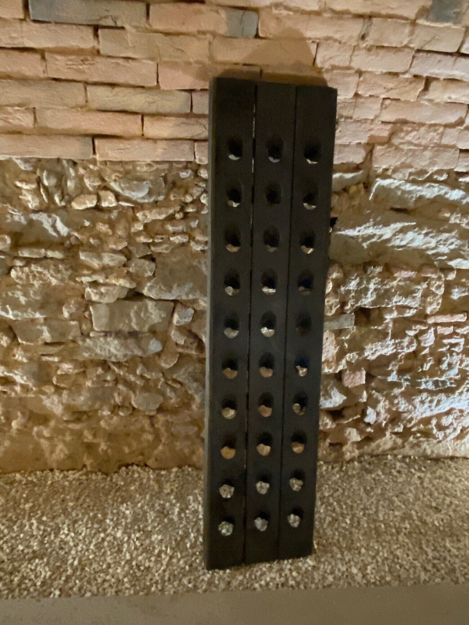 A black metal object with multiple holes against a stone wall.