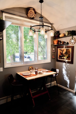 A dining area with a table set for two, a chandelier, and a window with a view of greenery and a street.