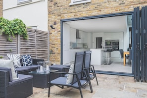 An outdoor patio with wicker furniture, a glass table, and a view into a modern kitchen and dining area.