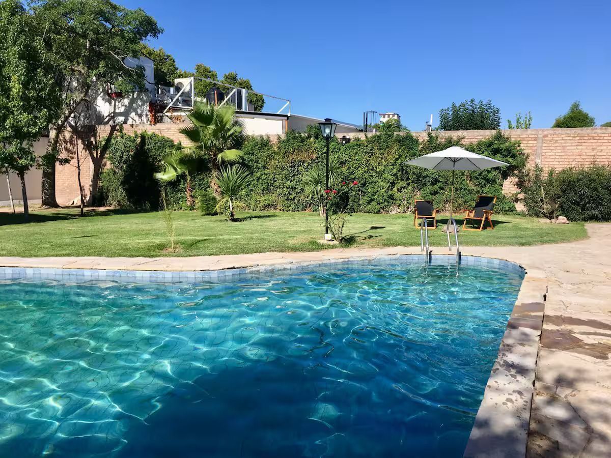 Una piscina con agua cristalina azul, rodeada de césped, árboles y un patio con sillas y una sombrilla.