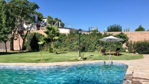 Una piscina con acqua cristallina, circondata da un'area verde con alberi e un patio con sedie e un ombrellone.
