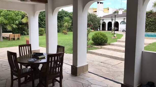 Una zona patio coperta con tavolo e sedie, una piscina e vista su un edificio con archi.