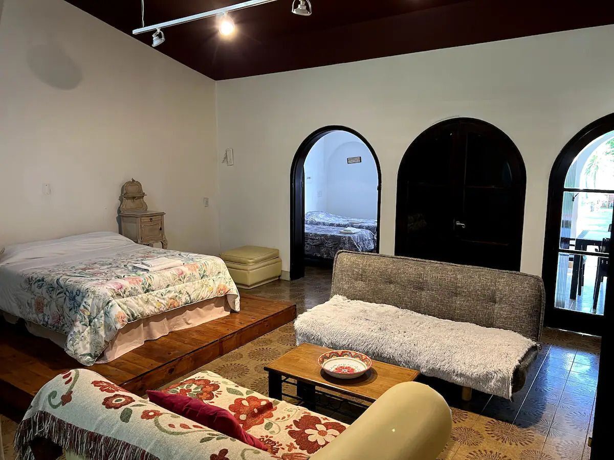Habitación con cama, sofá, una mesita y un banco. Cuenta con arcos en las puertas y vistas al exterior.