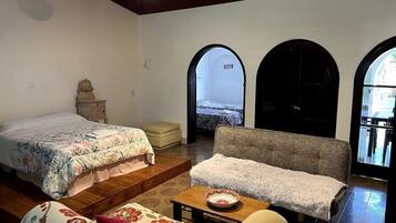 Habitación con cama, sofá, una mesita y un banco. Cuenta con arcos en las puertas y vistas al exterior.