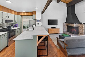 A modern kitchen with a central island, a stone fireplace, and a flat-screen TV. - Tahoe Corner - 3 Minutes to Tahoe, 7 to Northstar - Pool, Pickleball, Fitness (Kings Beach)
