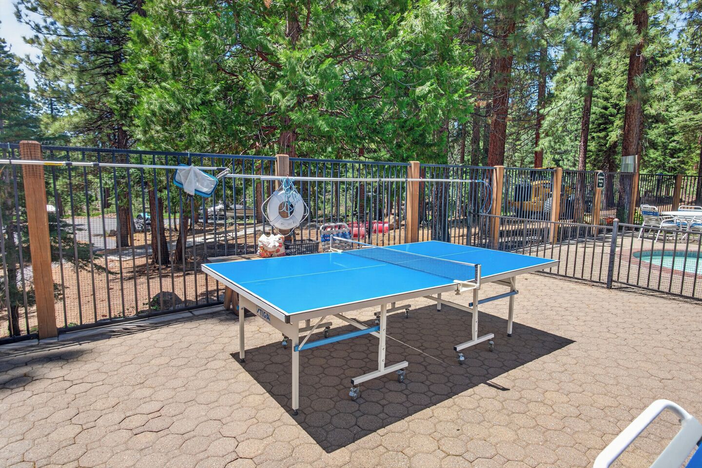 An outdoor ping pong table surrounded by a metal fence, with trees and a pool in the background.