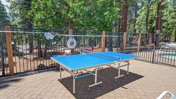 An outdoor ping pong table surrounded by a metal fence, with trees and a pool in the background.