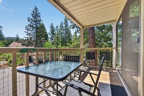 Eine überdachte Terrasse im Freien mit Tisch und Stühlen aus Glas, umgeben von einer Holzterrasse und Blick auf Bäume und Gebäude.