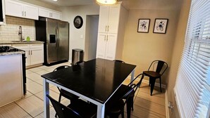 A compact kitchen with white cabinets, a stainless steel refrigerator, a black table, and a single chair.