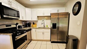 A kitchen with white cabinets, a stainless steel refrigerator, a microwave, a stove, and a trash can.