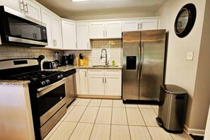 A kitchen with white cabinets, a stainless steel refrigerator, a microwave, a stove, and a trash can. - Decatur Executive Residential Stay | Sleeps 11 (Atlanta)