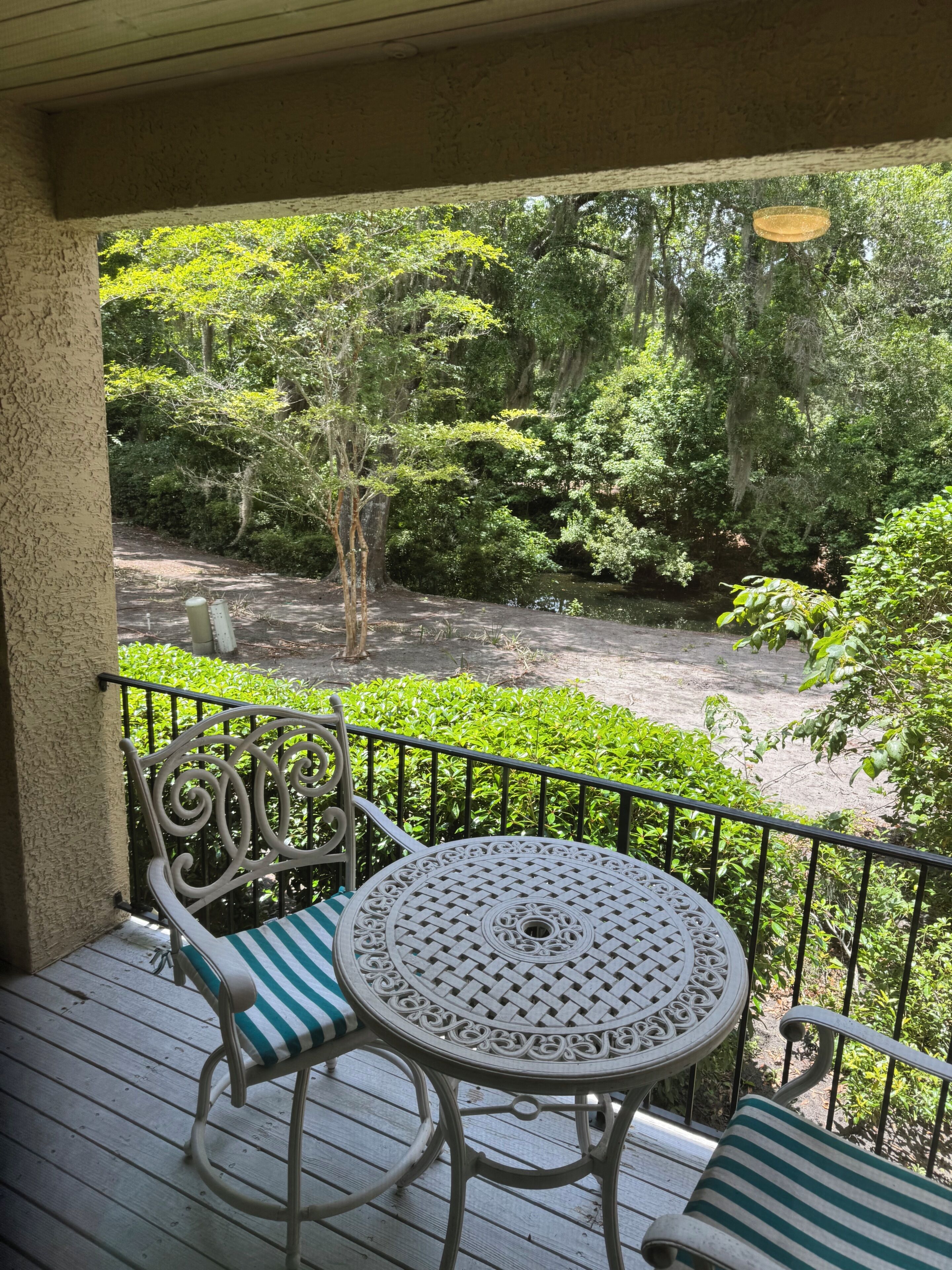 A balcony with a table and chairs, overlooking a garden with trees and a water feature.