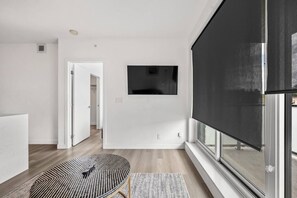 Ein modernes Wohnzimmer mit einem flachbildfernseher an der Wand, einem runden Tisch und einem Teppich mit Muster, sowie einem großen Fenster mit einer schwarzen Rollos.