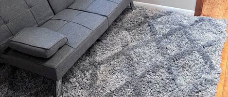 A grey sofa with a grey shaggy rug, a white wall, and a wooden floor.