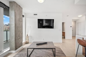 Ein modernes Wohnzimmer mit einem großen Fernseher, einer Betonstütze, einer Vase mit Zweigen und Blick auf die Stadt durch eine Glastür.