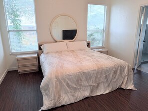 A bedroom with a bed, a bedside table, a nightstand, a round mirror, and two windows with blinds. - Awesome Renovated 3-bedroom house near Dallas Stadium (Euless)