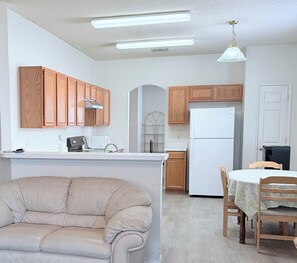 A compact kitchen with wooden cabinets, a white refrigerator, a dining table with chairs, and a sofa in the living area. - Awesome Renovated 3-bedroom house near Dallas Stadium (Euless)