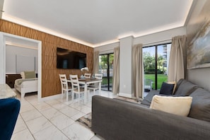 A modern living room with a sofa, a TV mounted on the wall, a dining area with chairs and a table, and a large window with curtains. - Studio Near Universal Lake View Pool & Gym (Orlando)