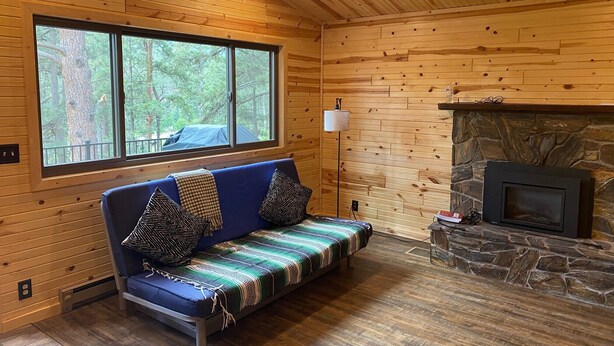 A cozy cabin interior with a blue sofa, a stone fireplace, and a large window overlooking a forest. - Gorgeous house for rent during Sturgis Rally 2026 (Rapid City)
