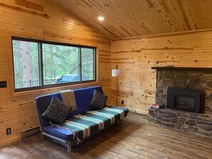 A cozy cabin interior with a blue sofa, a stone fireplace, and a large window overlooking a forest. - Gorgeous house for rent during Sturgis Rally 2026 (Rapid City)