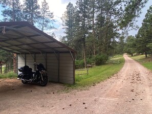 A covered motorcycle parking area adjacent to a gravel road, surrounded by trees. - Gorgeous house for rent during Sturgis Rally 2026 (Rapid City)