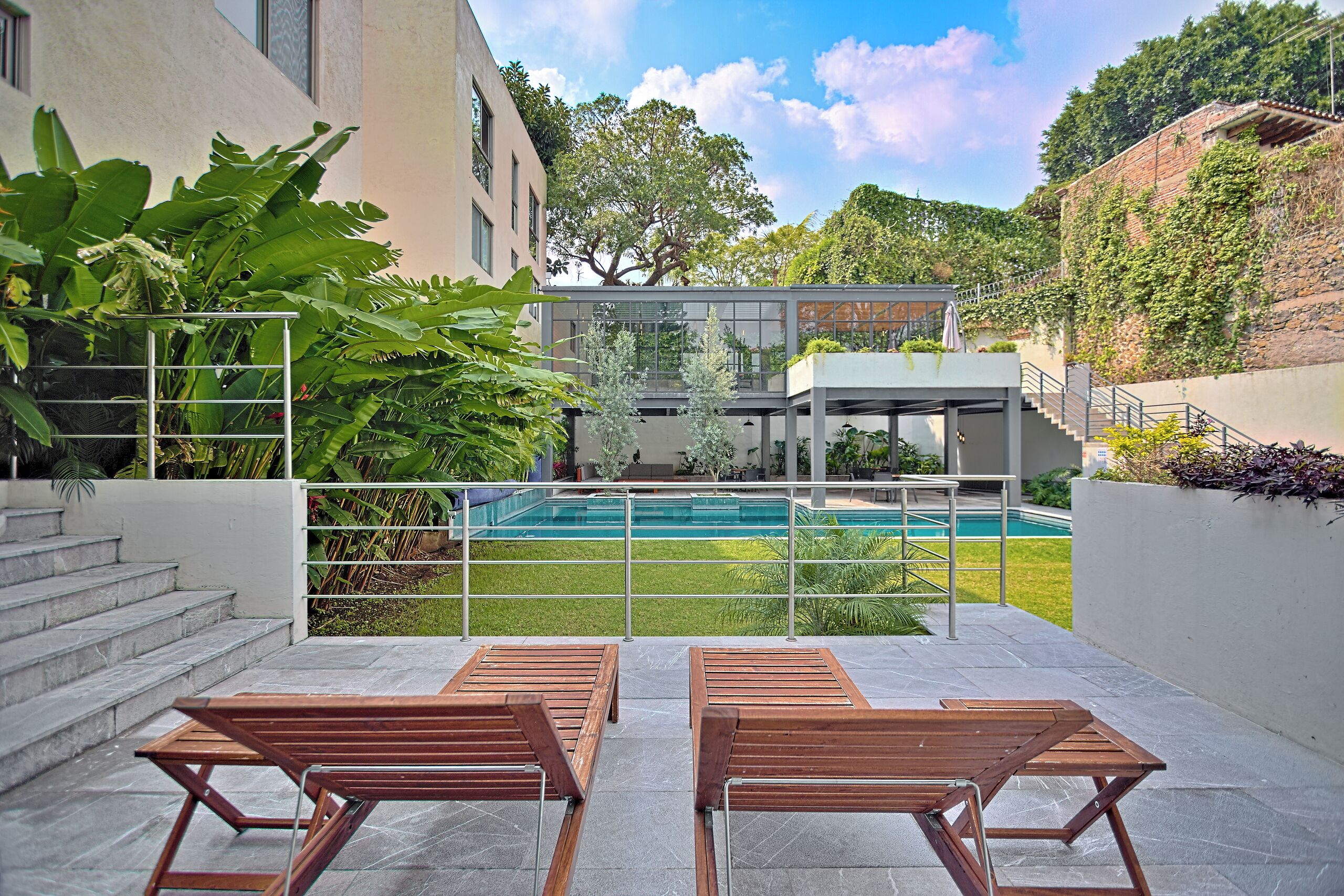 A modern outdoor area with a pool, wooden lounge chairs, and a staircase leading to a covered structure.