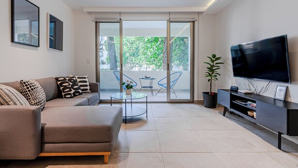 Un salon moderne avec un canapé, un téléviseur, une table en verre et un balcon équipé de chaises.