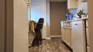 Una cocina estrecha con suelo de madera, armarios blancos y una zona de comedor con una silla.