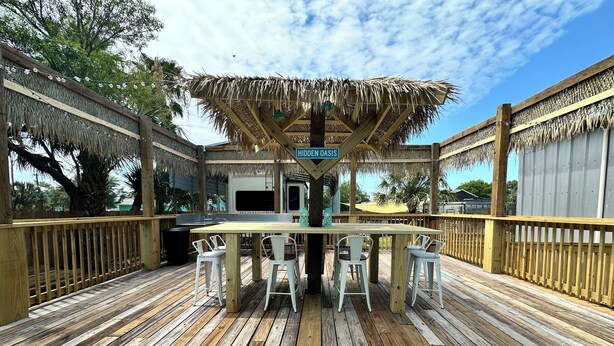 Una terrazza in legno con un'area bar ricoperta da un tetto di paglia, sedie bianche e un cartello con la scritta 'HIDDEN OASIS'.
