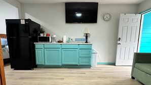Un soggiorno con ventilatore a soffitto, TV fissata al muro, angolo cottura con mobili color petrolio, frigorifero nero e tavolino in legno con telecomando.