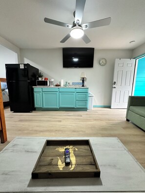 A living room with a ceiling fan, a TV mounted on the wall, a kitchen area with teal cabinets, a black refrigerator, and a wooden coffee table with a remote control. - Hidden Oasis Bunkhouse (Port O'Connor)