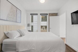 Un dormitorio con cama, mesita de noche, lámpara, televisor montado en la pared y ventana.