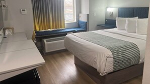 Ett hotellrum med en säng, ett skrivbord, ett fönster med gardiner, en värmare och ett nattduksbord med en lampa.
