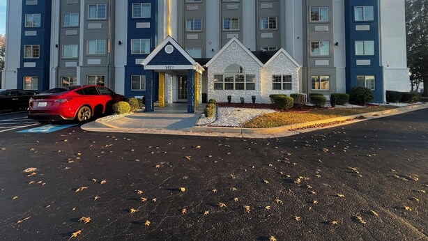 Ett hotell med flera våningar, en central entré, en parkeringsplats och en röd sportbil parkerad framför.