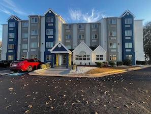 Et hotel med flere etager, en central indgang, en parkeringsplads og en rød sportsvogn foran.