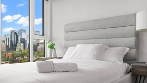Una habitación de hotel moderna con una cama grande, cabecera gris, mesita de noche con lámpara y vistas a la silueta urbana a través de un ventanal.