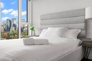 Ein modernes Hotelzimmer mit einem großen Bett, einem grauen Kopfteil, einem Nachttisch mit Lampe und Blick auf die Stadtkulisse durch ein großes Fenster.