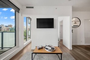 Ein modernes Wohnzimmer mit großem Fenster, einem wandmontierten Flachbildfernseher, einem Holztisch und Blick auf die Stadtlandschaft.
