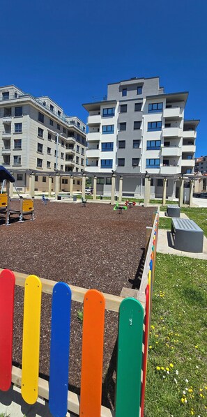 A playground with a wooden fence, a parking area, and a multi-story residential building in the background.