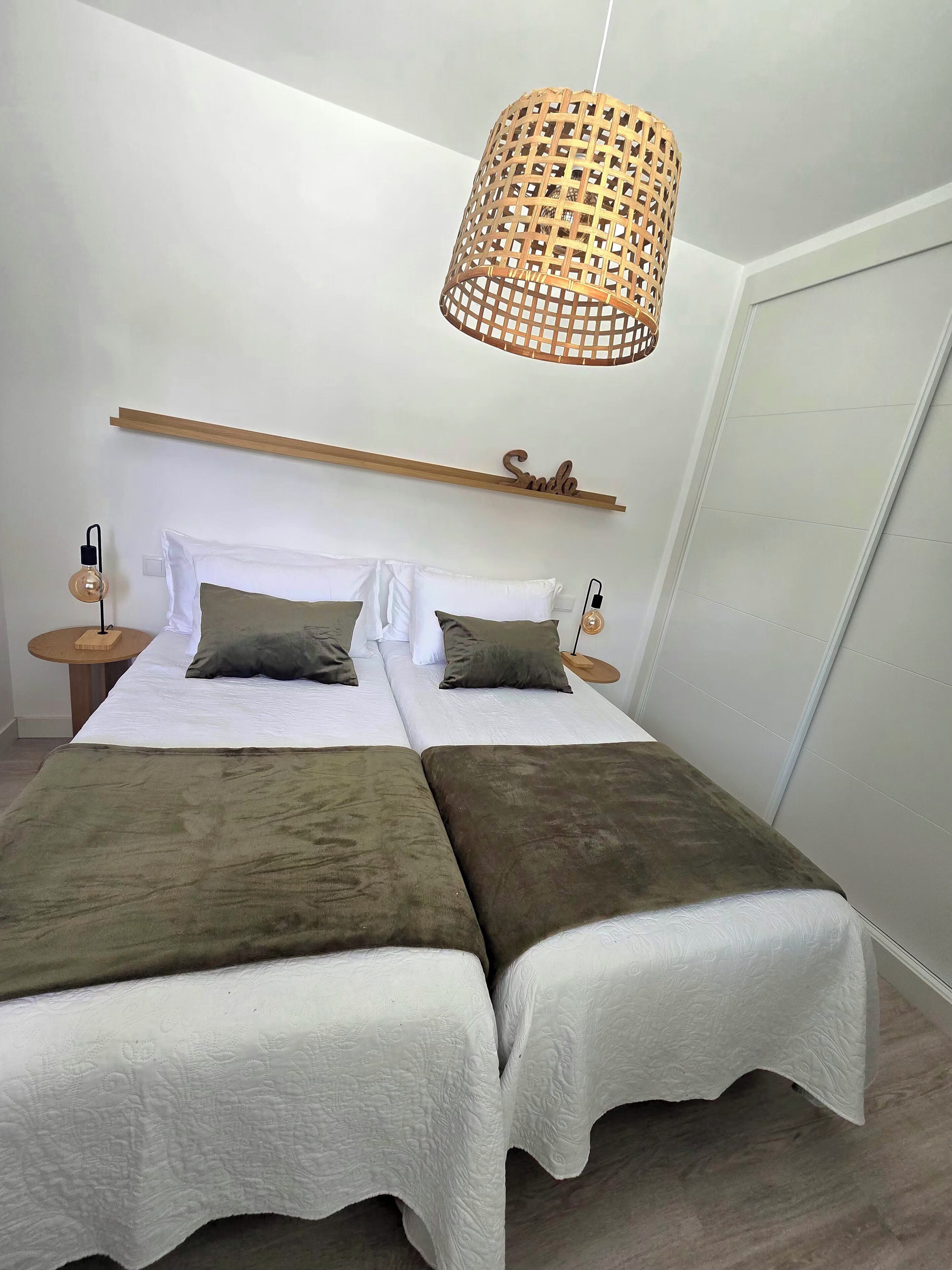 A double bed with two pillows, a bedside table with a lamp, a wooden shelf with a 'Smile' sign, and a woven light fixture.