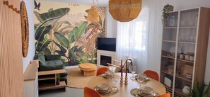 A modern living room with a large tropical mural, a dining area with a round table and orange chairs, a television, and a bookshelf.