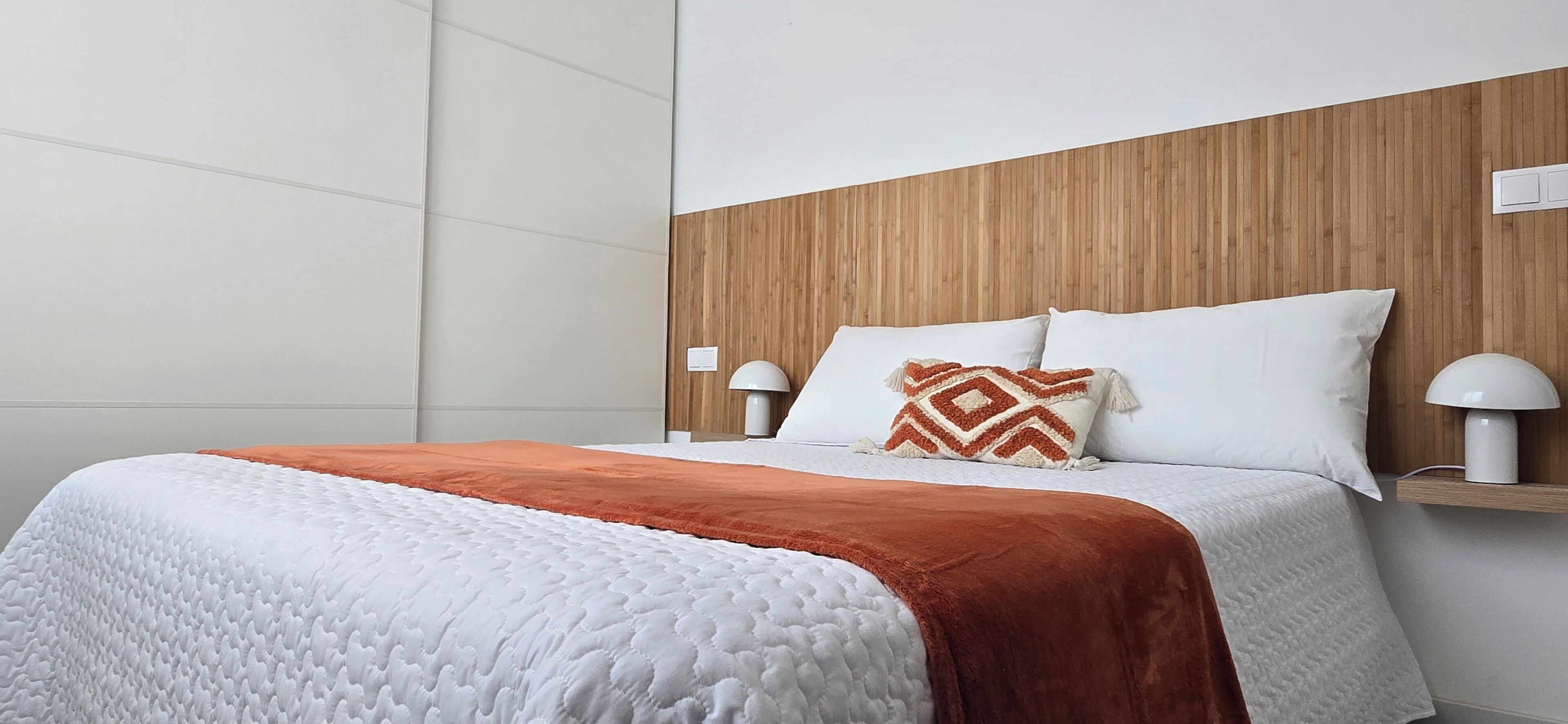 A neatly made bed with a white and orange blanket, two pillows, and a wooden headboard.