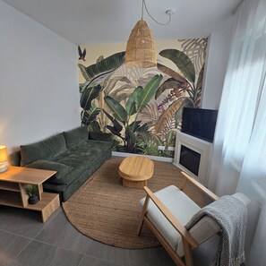 Ein Wohnzimmer mit einem grünen Sofa, einem runden Holztisch, einem Sessel, einem Teppich und einer großen Wandmalerei mit tropischen Pflanzen.