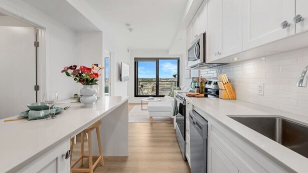 A modern kitchen with white cabinetry, a central island, and a view to the outdoors. - NEW | Mins to Saddledome | Rooftop Patio | UG Park (Calgary)