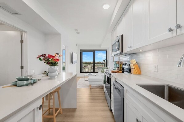 A modern kitchen with white cabinetry, a central island, and a view to the outdoors. - NEW | Mins to Saddledome | Rooftop Patio | UG Park (Calgary)