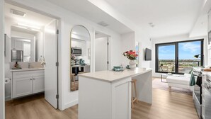 A modern kitchen with white cabinetry, a central island, and a large window offering a city view. - NEW | Mins to Saddledome | Rooftop Patio | UG Park (Calgary)