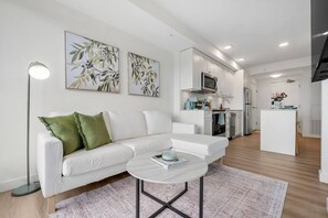 A modern living room with a white sofa, a round coffee table, and a kitchen in the background. - NEW | Mins to Saddledome | Rooftop Patio | UG Park (Calgary)